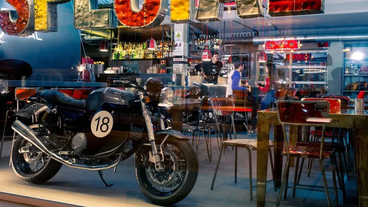 Un bar per motociclisti con una moto d'epoca in primo piano. Dietro, il bancone &egrave; pieno di bottiglie e personale al lavoro.
