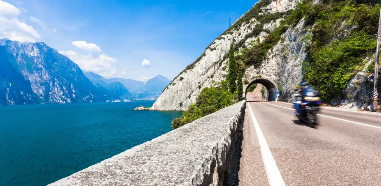 Un motociclista sfreccia lungo la strada panoramica del Lago di Garda, con montagne imponenti e il blu intenso dell'acqua.