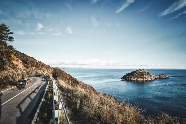 Un motociclista percorre una strada panoramica sull'Argentario, con il mare blu e un'isola rocciosa sullo sfondo.