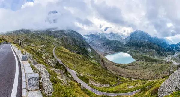 Strada tortuosa sul passo della Sambuca, con lago alpino azzurro e montagne innevate sullo sfondo.